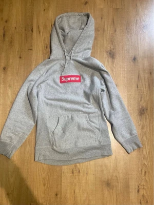 Grå Supreme hoodie med box logo - Snygg grå hoodie från Supreme med klassisk röd box logo på bröstet. Hoodien har huva med dragsko och en stor magficka framtill. Tillverkad i mjukt bomullsmaterial och har ribbade muddar vid ärmslut och nederkant. Perfekt för en streetwear-look.