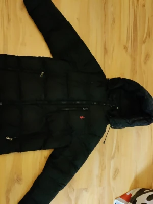 Svart pufferjacka från Polo Ralph Lauren - Svart pufferjacka från Polo Ralph Lauren med huva och dragkedja framtill. Jackan har puffad design, två sidofickor med dragkedja och Polo-logga i rött på bröstet. Perfekt för kalla dagar och har en riktigt schysst streetstil.