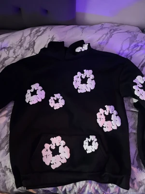 Svart hoodie med vita blommor - Snygg svart hoodie med stora vita blommönster tryckta över hela tröjan, även på ärmar och ficka. Hoodien har en klassisk huva och magficka framtill. Perfekt för dig som vill sticka ut med en unik och trendig stil. Materialet känns mjukt och bekvämt.