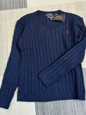 Mörkblå kabelstickad tröja Polo Ralph Lauren - Mörkblå kabelstickad tröja från Polo Ralph Lauren med röd broderad logga på bröstet. Tröjan har lång ärm, ribbstickade muddar och klassisk v-ringning på ena och rund hals på andra. Perfekt för en stilren och avslappnad look.