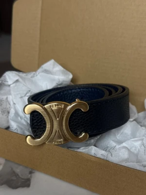 Svart bälte från Celine med guldspänne - Stilrent svart bälte från Celine i präglat skinn med ikoniskt guldigt Triomphe-spänne. Perfekt accessoar för att lyfta din outfit med en lyxig touch och klassisk design.