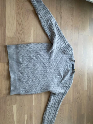 Ljusgrå kabelstickad tröja Polo Ralph Lauren - Ljusgrå kabelstickad tröja från Polo Ralph Lauren med rund halsringning och diskret broderad logga på bröstet. Tröjan har långa ärmar och ribbade muddar vid ärmslut och nederkant. Perfekt för dig som gillar klassisk och stilren look.
