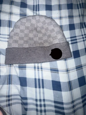 Grå rutig mössa med uppvikt kant - Snygg grå mössa med diskret rutmönster och bred uppvikt kant. Perfekt accessoar för kyliga dagar och passar till de flesta outfits. Tillverkad i mjukt material som känns skönt mot huden.