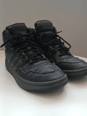 Svarta Adidas kängor med skön fodring - Köpte dom men tyvärr så passade dom inte. Säljer ett par svarta Adidas kängor med klassiska tre ränder på sidan och högt skaft. Skorna har snörning, vadderad plös och krage samt mjukt foder för extra komfort. 