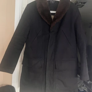 Svart rock med fuskpäls från Filippa K - Stilren svart rock från Filippa K med bred krage i brun fuskpäls. Klassisk rak passform, två stora fickor framtill och diskreta sömdetaljer. Perfekt för dig som vill ha en tidlös och snygg look under kallare dagar.