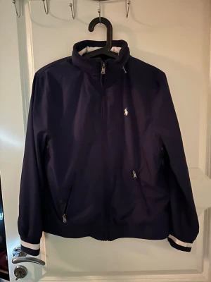 Marinblå vindjacka från Polo Ralph Lauren - Snygg marinblå vindjacka från Polo Ralph Lauren med vit logga på bröstet. Jackan har dragkedja framtill, två fickor med dragkedja och vita detaljer på muddarna. Hög krage och sportig look, perfekt för blåsiga dagar.passar dig mellan 155 till 160