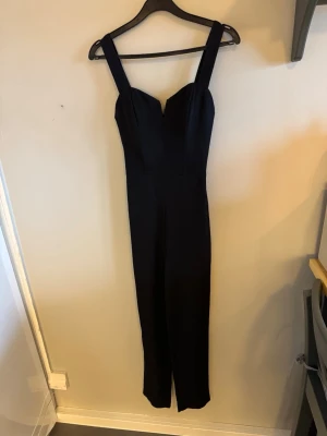 Svart jumpsuit med breda axelband - Snygg svart jumpsuit med breda axelband och figurnära överdel. Byxdelen är rak och lång, vilket ger en clean och stilren look. Perfekt för dig som vill ha något enkelt men ändå trendigt. Materialet ser ut att vara mjukt och följsamt, troligen polyester eller bomullsblandning.