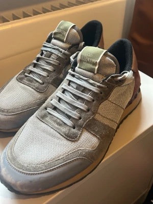 Grå sneakers från Valentino - Snygga sneakers från Valentino i grått med mesh och mockadetaljer. Skorna har svarta och mörk röda inslag samt en ljusgrön logga på plösen. Klassisk låg modell med vita och grå skosnören och svart innersula.