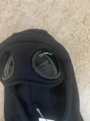 C.P. Company Goggle Hoodie – Svart – S – Ny med tags - Snabb affär kom privat! Säljer en svart hoodie från C.P. Company med dragkedja och ikoniska inbyggda glasögon i huvan. Hoodien har två stora fickor framtill och ribbade muddar. Perfekt för dig som vill sticka ut med en unik streetwear-look.
