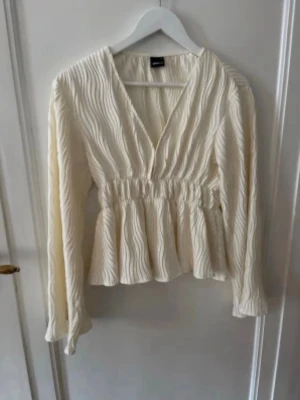 Krämvit blus med struktur - Snygg krämvit blus från Gina Tricot med vågig struktur i tyget. Djup v-ringning, markerad midja och volang nertill. Långa ärmar och lätt, luftig känsla. Perfekt att styla med jeans eller kjol.