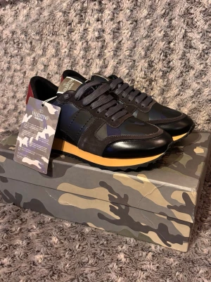 Valentino Rockrunner - Snygga svarta sneakers från Valentino Garavani med detaljer i mörkblått och vinrött. Skorna har en gul sula och är gjorda i en mix av läder och textil. Klassisk låg modell med svarta skosnören och coola camo-detaljer. Perfekt för dig som vill ha något extra till din outfit. Og följer med, och de är aldrig använda