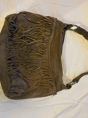 Brun mocka axelväska med fransar - Säljer en snygg brun axelväska i mocka med långa fransar på framsidan. Väskan har en bred axelrem med metallringar och dragkedja upptill. Perfekt för dig som gillar boho-stil och vill ha en unik accessoar. Har lite missfärgning på dragkedjan och några fläckar och några fransar har åkt av men man tänker inte så mycket på det