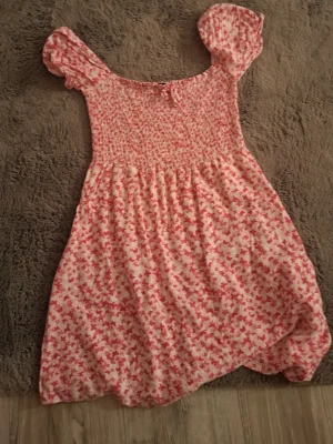 Rosa blommig kortklänning med puffärm - Supersöt kortklänning med rosa blommigt mönster på vit botten. Klänningen har smock upptill, puffärmar och en liten rosett framtill. Perfekt för sommarens alla tillfällen och ger en romantisk vibe.