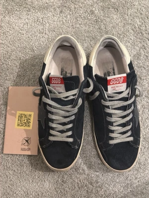 Svarta Golden Goose sneakers - Snygga mörkblå sneakers från Golden Goose med vita detaljer och grå skosnören. Skorna har en klassisk låg modell med vit sula och GGDB-logga på plösen. Tillverkade i mocka och läder för en exklusiv känsla.