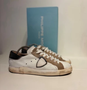 Philippe Model sneakers i vitt och beige - Snygga Philippe Model sneakers i vitt skinn med beige och svarta detaljer. Klassisk låg modell med rund tå, platt sula och snörning. Ikonisk sköldlogga på sidan och kontrastfärgad häl. Perfekta för en trendig streetstyle-look.