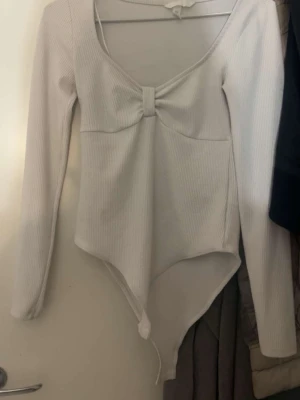 Vit ribbad body med rosett - Snygg vit body i ribbat material med lång ärm och en söt rosettdetalj framtill. Djup ringning och klassisk knäppning nedtill. Perfekt att styla till jeans eller kjol för en trendig look.