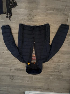 Mörkblå dunjacka med huva från Parajumpers - Snygg mörkblå dunjacka från Parajumpers med huva och två dragkedjeförsedda fickor framtill. Jackan har quiltad design och gul detalj vid halsen. Perfekt för kalla vinterdagar och riktigt skön att bära.