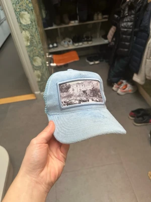 Ljusblå truckerkeps med tryck - Säljer en ljusblå truckerkeps med mesh baktill och justerbar snapback. Framtill finns ett svartvitt tryck med motiv av en byggnad och folkmassa. Perfekt för dig som vill ha en cool och avslappnad stil.