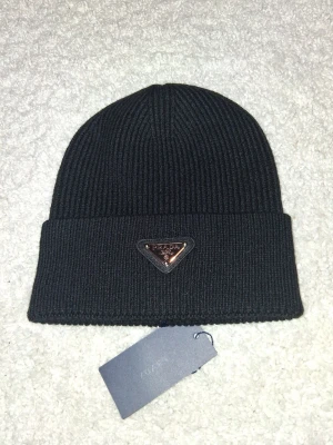 Svart ribbstickad mössa från Prada - Svart ribbstickad mössa från Prada med klassisk uppvikt kant och metalltriangel med logga framtill. Snygg och stilren design som passar perfekt till kalla dagar. Mössan är tillverkad i mjukt material och har en exklusiv känsla.