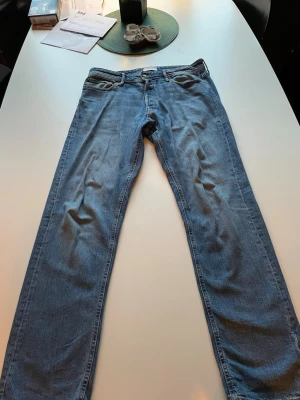 Blå raka jeans med klassisk passform - Säljer ett par blå jeans med rak passform och fem fickor. Jeansen har en klassisk look med normal midja och dragkedja. Materialet är denim i bomull och färgen är ljusblå. Perfekta för en avslappnad stil.
