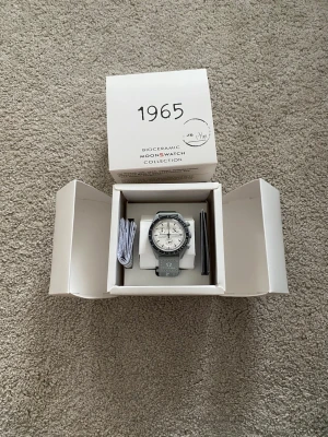 Omega x Swatch MoonSwatch klocka - Stilren Omega x Swatch MoonSwatch från Bioceramic-kollektionen. Klockan har en grå boett, vit urtavla med svarta detaljer och grått armband med Omega-logga. Kommer i originalförpackning med tillhörande manualer.