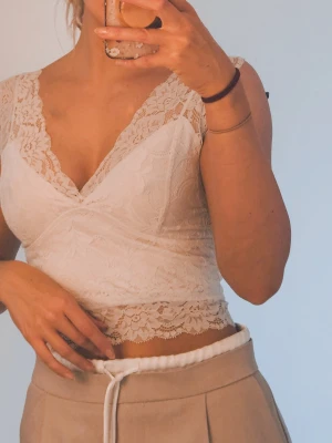 Vit spetsblus med v-ringning - Superfin vit blus i spets med v-ringning både fram och bak. Blusen är ärmlös och har ett blommigt spetsmönster över hela plagget. Perfekt för dig som gillar romantiska detaljer och vill ha något som sticker ut lite extra. Vissa bilder såg den gul ut mot banden men har tagit en närbild så man förstår att det bara är ljuset som gör att det ser ut så! 
