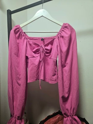 Rosa blus med snörning  - Säljer en rosa blus med långa puffärmar och smockad rygg. Toppen har en söt snörningsdetalj framtill och resår vid axlar och ärmslut. Mycket stretch tack vare resåren i ryggen! Bild från samma I svart