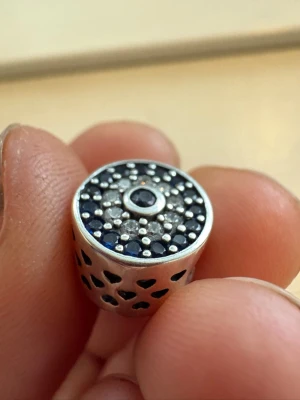 Silverfärgad pärla med blå stenar - Snygg rund pärla i silverfärgad metall med blå och klara stenar på ovansidan. Sidorna har utskurna hjärtan som ger en extra detalj. Perfekt att trä på ett armband för en personlig touch.