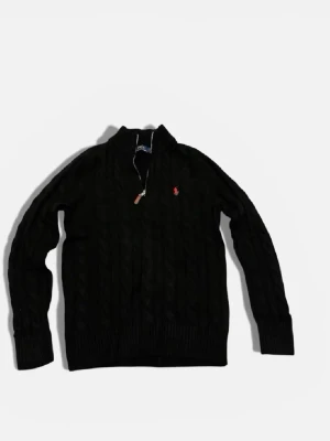 Svart kabelstickad tröja Polo Ralph Lauren - Kabelstickad svart tröja från Polo Ralph Lauren med halv dragkedja och röd broderad logga på bröstet. Tröjan har hög krage, långa ärmar och ribbade muddar. Perfekt för dig som gillar klassisk och stilren look.