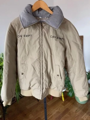 Beige bomberjacka från Cav Empt - Snygg beige bomberjacka från Cav Empt med svart broderad logga på bröstet. Jackan har en stor grå krage i mjukt material, dragkedja framtill och två fickor med knapp. Perfekt för dig som gillar streetwear och vill sticka ut.
