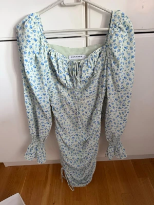 Blommig långklänning från Adoore - Romantisk långklänning från Adoore med blått blommönster på ljus botten. Klänningen har långa ärmar med volang vid ärmslut, snörning vid halsringningen och figurnära passform. Perfekt för sommarens alla tillfällen.