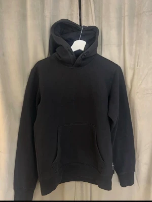 Svart hoodie från Sail Racing - Klassisk svart hoodie från Sail Racing med stor känguruficka framtill och ribbade muddar vid ärmar och nederkant. Hoodien har en rejäl huva med dragsko och är tillverkad i mjukt bomullsmaterial. Perfekt för en avslappnad och stilren look.