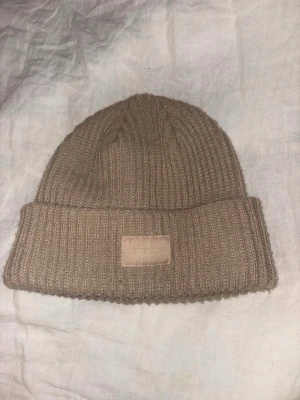 Beige ribbstickad mössa  - Snygg beige ribbstickad mössa med uppvikt kant och diskret logopatch framtill. Tillverkad i mjukt och värmande material, perfekt för kyliga dagar. Enkel och stilren design som passar till det mesta.