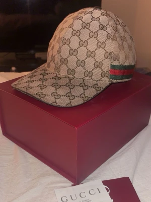 Gucci beige GG canvas keps - Säljer en beige keps från Gucci i klassisk GG canvasmönster. Kepsen har böjd skärm och detaljer med grönt och rött band på sidan. Tillverkad i canvasmaterial och har en exklusiv känsla som passar perfekt till streetwear eller för att lyfta din outfit.