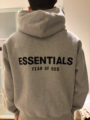 Dark oatmeal Essentials Fear of God hoodie - Säljer en grå hoodie från Essentials Fear of God i storlek xs med stor logga på ryggen och mindre logga på bröstet. Hoodien har huva och en stor magficka framtill. Tillverkad i mjukt sweatshirtmaterial, perfekt för en chill och stilren look.
