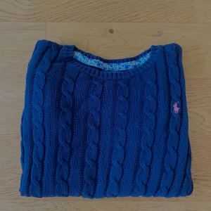 Marinblå kabelstickad tröja Polo Ralph Lauren - Kabelstickad marinblå tröja från Polo Ralph Lauren med klassisk rund hals och lång ärm. Tröjan har ett broderat rosa Polo-logo på bröstet och ribbade muddar vid ärmslut och nederkant. Snabb affär 