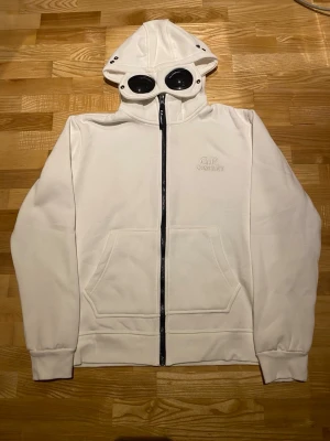 Vit hoodie med goggles från C.P. Company - Säljer en vit hoodie från C.P. Company med ikoniska goggles i huvan och dragkedja framtill. Tröjan har känguruficka, ribbade muddar och broderad logga på bröstet. Perfekt för dig som gillar streetwear och unika detaljer.