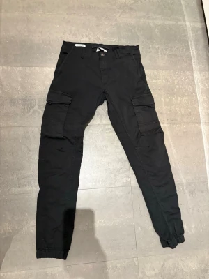 Svarta cargobyxor med resår - Svarta cargobyxor med flera stora fickor på sidorna och resår vid bensluten. Byxorna har en avslappnad passform och stängs med knapp och dragkedja. Perfekta för en streetwear-look och praktiska med sina många fickor.