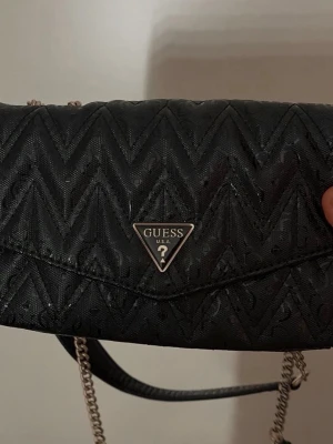 Svart axelväska från Guess - Snygg svart axelväska från Guess med kedjerem och quiltat mönster i zigzag. Väskan har ett metallfärgat Guess-emblem framtill och dragkedja på baksidan. Materialet är konstläder med glansig finish. Perfekt för att lyfta din outfit.