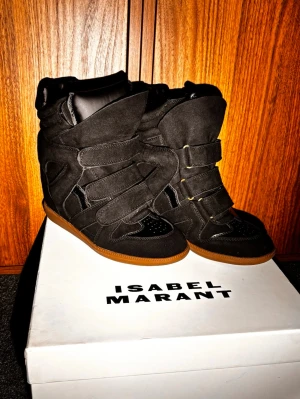 Svarta Isabel Marant sneakers - Köpte dessa till min tjej inför alla hjärtans dag men tyvärr kommer hon ej få dessa längre. Snygga svarta sneakers från Isabel Marant med högt skaft och kilklack. Skorna har flera kardborreband, detaljer i mocka och läder samt en brun gummisula. Perfekta för dig som vill ha en edgy och trendig look.