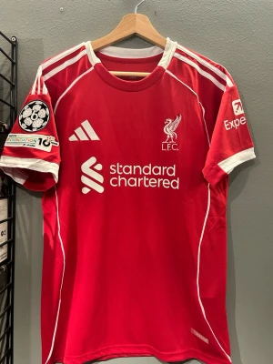 Liverpool matchtröja röd Adidas - Röd Liverpool FC matchtröja från Adidas med vita detaljer, klubbmärke och sponsortryck framtill. Nummer 22 och namnet Ekitike på ryggen. Champions League-märke på ärmen och vita ränder på axlarna. Perfekt för fotbollsfans! Helt ny med tagg en replika 