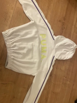 Vit hoodie från Puma med lila detaljer - Snygg vit hoodie från Puma med lila ränder längs ärmarna och stort PUMA-tryck i gult på ryggen. Hoodien har huva och resår i midjan för en skön passform. Perfekt för dig som gillar sportig och avslappnad stil.