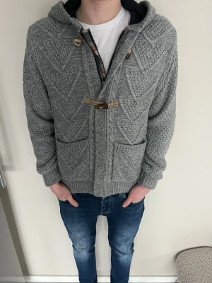 Grå stickad cardigan  - Riktigt fet grå stickad cardigan med träknappar och två framfickor. Tröjan har ett tydligt mönster i stickningen och ribbade muddar vid ärmslut och nederkant. Perfekt för lager på lager och chill vardagsstil. Modellen är 175 och väger 60kg 