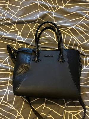 Svart handväska från Michael Kors - Snygg svart handväska från Michael Kors med både släta och ormmönstrade detaljer. Väskan har två handtag, axelrem och guldfärgad logga framtill. Tillverkad i konstläder och har en stilren, strukturerad form.