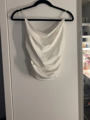 Vit satinliknande linnetopp - Säljer en vit linnetopp med tunna axelband och draperad framsida. Toppen har en slät baksida och ett glansigt, satinliknande material som ger en lyxig känsla. Perfekt att styla med jeans eller kjol för en trendig look. Finns ingen lapp i den så har ingen aning om vilket märke eller storlek 