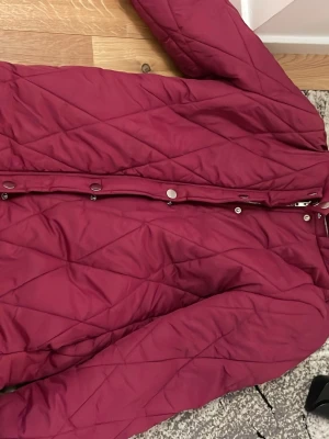 Vinröd pufferjacka med knappar - Säljer en vinröd pufferjacka med snyggt quiltat mönster och dragkedja framtill. Jackan har tryckknappar längs dragkedjan och är vadderad för extra värme. Perfekt för kalla dagar och riktigt skön att bära.