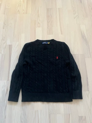 Svart kabelstickad tröja Polo Ralph Lauren - Kabelstickad svart tröja från Polo Ralph Lauren med rund halsringning och röd broderad logga på bröstet. Tröjan har långa ärmar och ribbade muddar vid ärmslut och nederkant. Perfekt för en stilren och klassisk look.