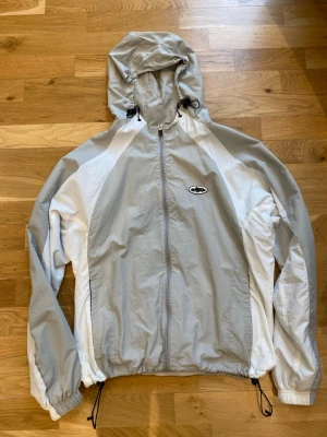 Ljusgrå och vit Corteiz vindjacka  - Snygg vindjacka från Corteiz i ljusgrått och vitt med dragkedja framtill, justerbar huva och elastiska muddar. Jackan har logotyp på bröstet och dragsko i nederkant. Perfekt för blåsiga dagar och streetwear-stil.