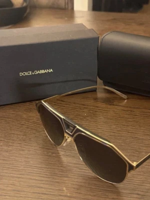 Guldiga solglasögon från Dolce & Gabbana - Snygga solglasögon från Dolce & Gabbana med guldig metallbåge och mörka glas. Modellen har en modern, kantig form och tunna skalmar med märkets logga på sidan. Perfekt accessoar för dig som vill sticka ut med en exklusiv look.
