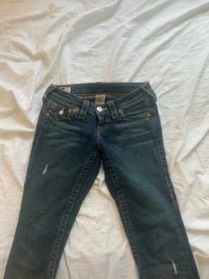 Mörkblå 2000s True Religion jeans - Snygga mörkblå jeans från True Religion med klassisk femficksdesign och diskreta slitningar. Jeansen har orangea kontrastsömmar och låg midja. Tillverkade i USA och har detaljerad insida med mönstrat tyg vid fickorna.